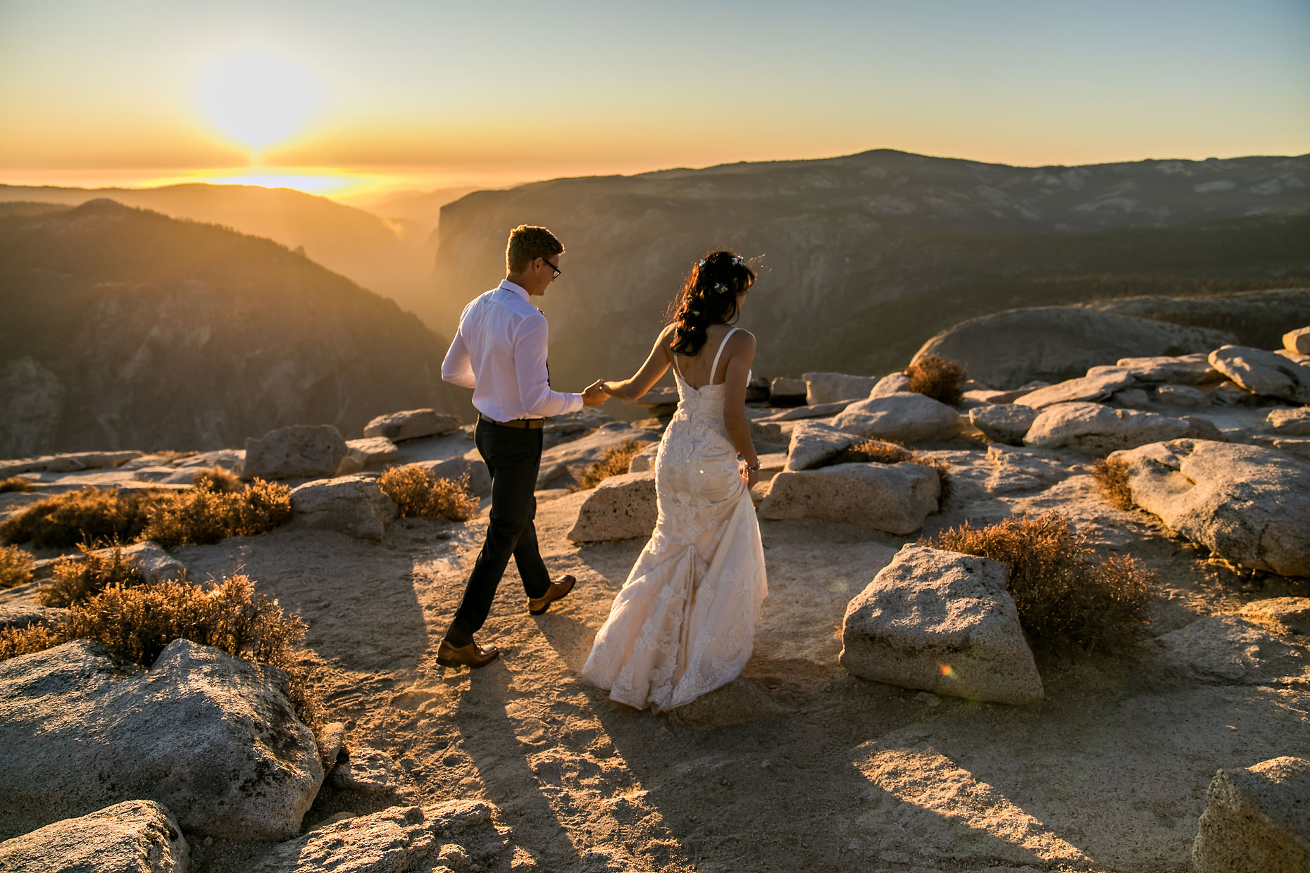Bride & groom in wedding garb walking hand in hand in Yosemite at sunset.